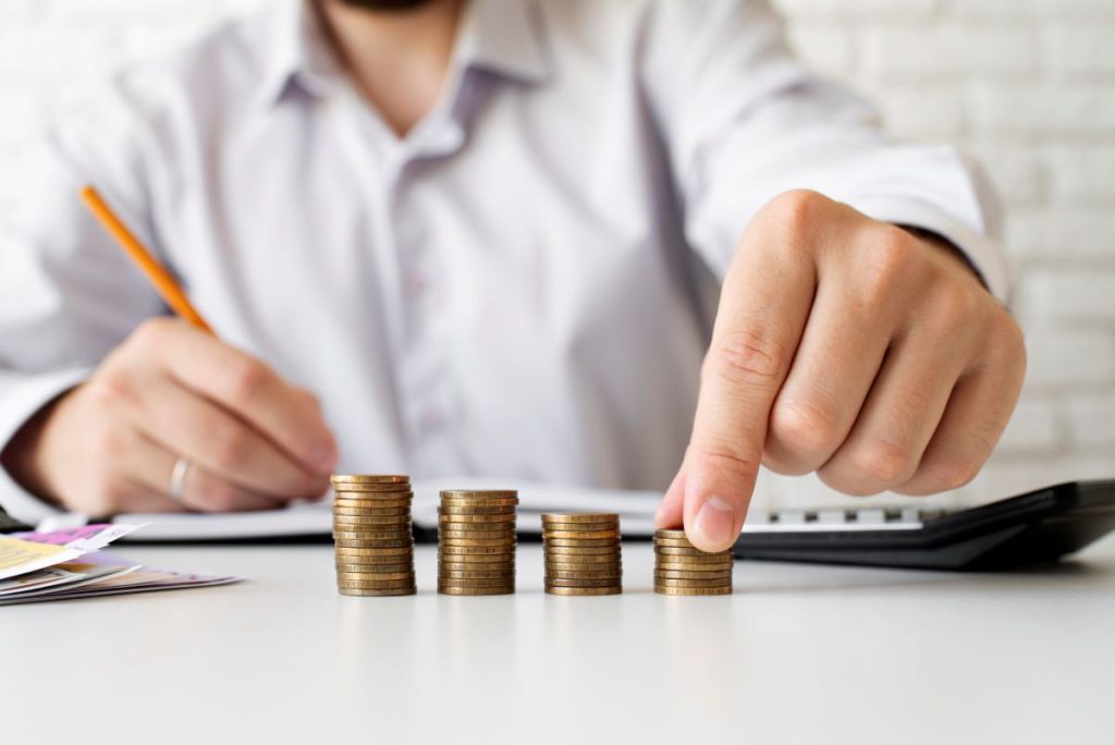Mężczyzna przy biurku notujący ołówkiem i układający stosy monet obok kalkulatora, co symbolizuje programy oszczędnościowe i zarządzanie finansami.