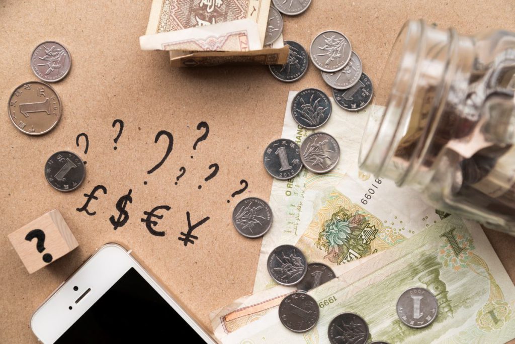 Banknoty i monety obok smartfona i narysowanych symboli walut ze znakami zapytania, co ilustruje planowanie budżetu i programy oszczędnościowe.